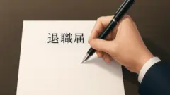 木目の濃いデスクの上に斜めに置かれた白い用紙の左寄りに退職届の文字があり、その右側で紺色のスーツの袖をした人の右手が黒い万年筆を握って書き出そうとしている様子