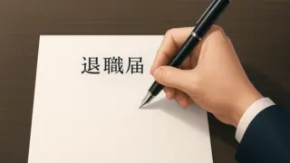 木目の濃いデスクの上に斜めに置かれた白い用紙の左寄りに退職届の文字があり、その右側で紺色のスーツの袖をした人の右手が黒い万年筆を握って書き出そうとしている様子