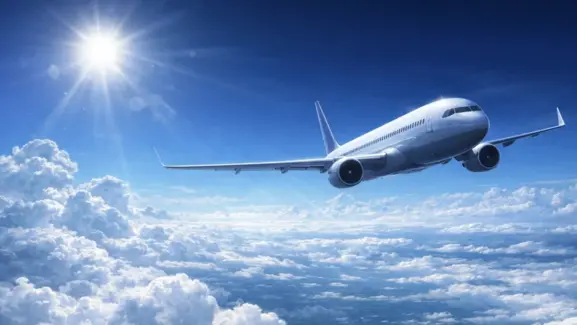 深い青空と強い日差しの下、雲海の上を白い旅客機が滑るように巡航し、光の筋とレンズフレアが広がる開放的な空撮風シーン