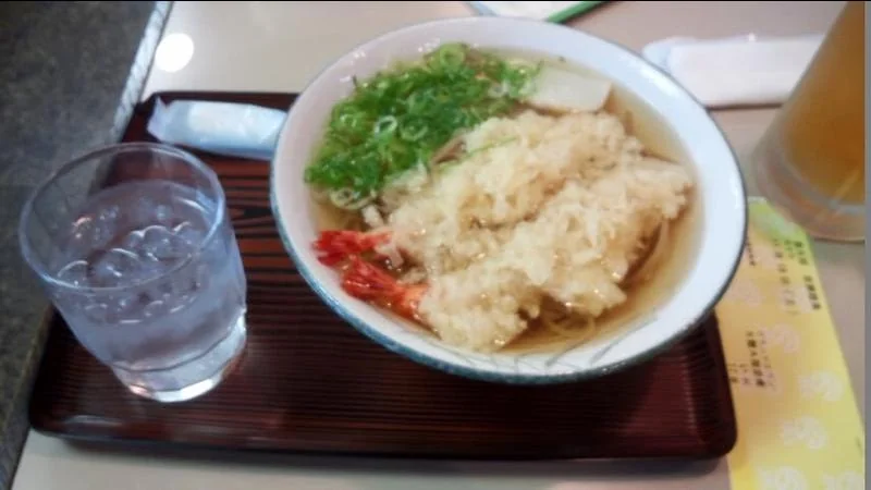 京都競馬場のA指定席エリアにあった「駒」の天ぷらそばで、黒いお盆の上にねぎと天ぷらが乗ったそばの丼が置かれ、横に水のグラスと奥にビールが見えるテーブル写真