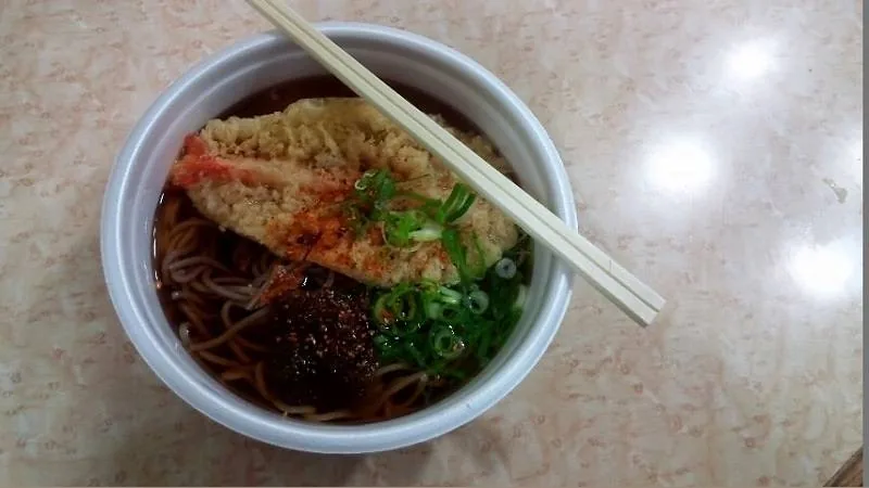 京都競馬場のレジャーフーズの天ぷらそばで、白い丼にだしの入ったそばと天ぷらと刻みねぎが乗り、ベージュのテーブルに割り箸を添えて置かれている俯瞰写真