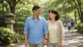 晴れた昼の日本の都市公園で、50代〜60代の日本人カップルが手をつないで並んで歩き、木漏れ日と緑に包まれた遊歩道の奥に人影がぼけて見え、石灯籠と竹柵がさりげなく写る爽やかな雰囲気