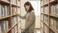 図書館の書架の間で女性が背表紙を指で選びカゴを持って歩く柔らかな自然光