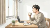 朝の光が差す部屋の木の机で、ノートパソコンとメモ帳の横に座り、マグカップを手に一息つく男性の横顔
