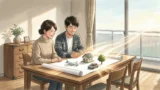 日本人夫婦がリビングの木製テーブルで家の模型と旅行パンフレットを広げて指さしている